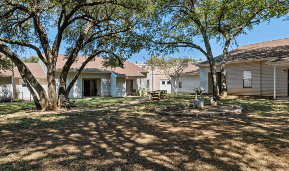 Sunny courtyard with large shade trees, benches, a picnic table and a small decorative fountain surrounded by single-story retirement community buildings.