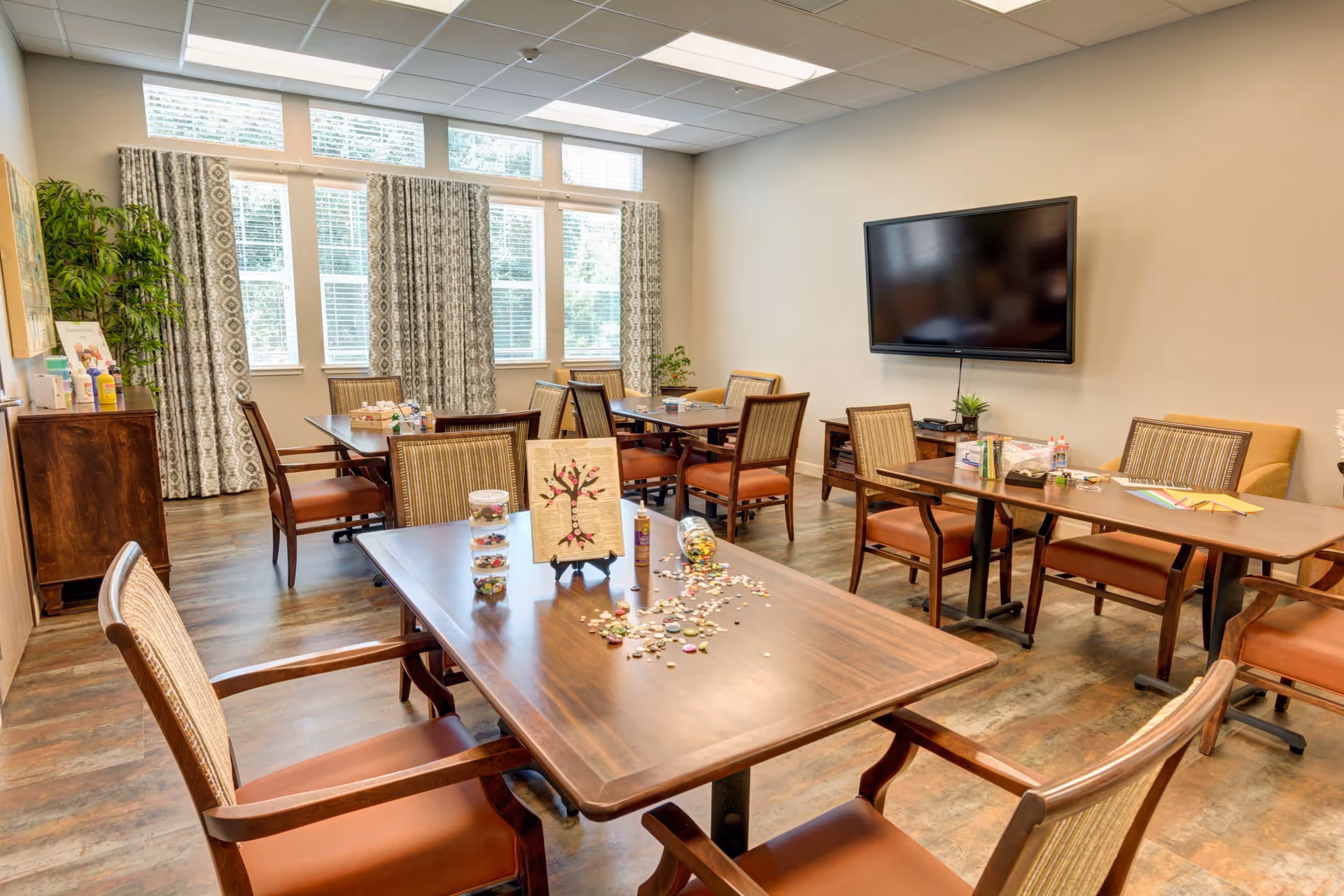 A bright room with multiple wooden tables and chairs arranged for group activities. The tables have craft supplies, including a small tree artwork, jars with colorful items, and scattered buttons or beads. Large windows with patterned curtains let in natural light. A flat-screen TV is mounted on the wall, and a wooden cabinet with plants and supplies is in the corner.
