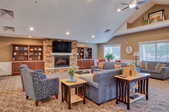 A spacious senior living common area with comfortable seating including two patterned armchairs and two gray sofas arranged around a stone fireplace with a mounted flat-screen TV above it. Built-in wooden shelves flank the fireplace, holding decorative items and books. The room has large windows with blinds, a ceiling fan with lights, and a carpeted floor. Side tables with plants and decorative items are placed near the seating.
