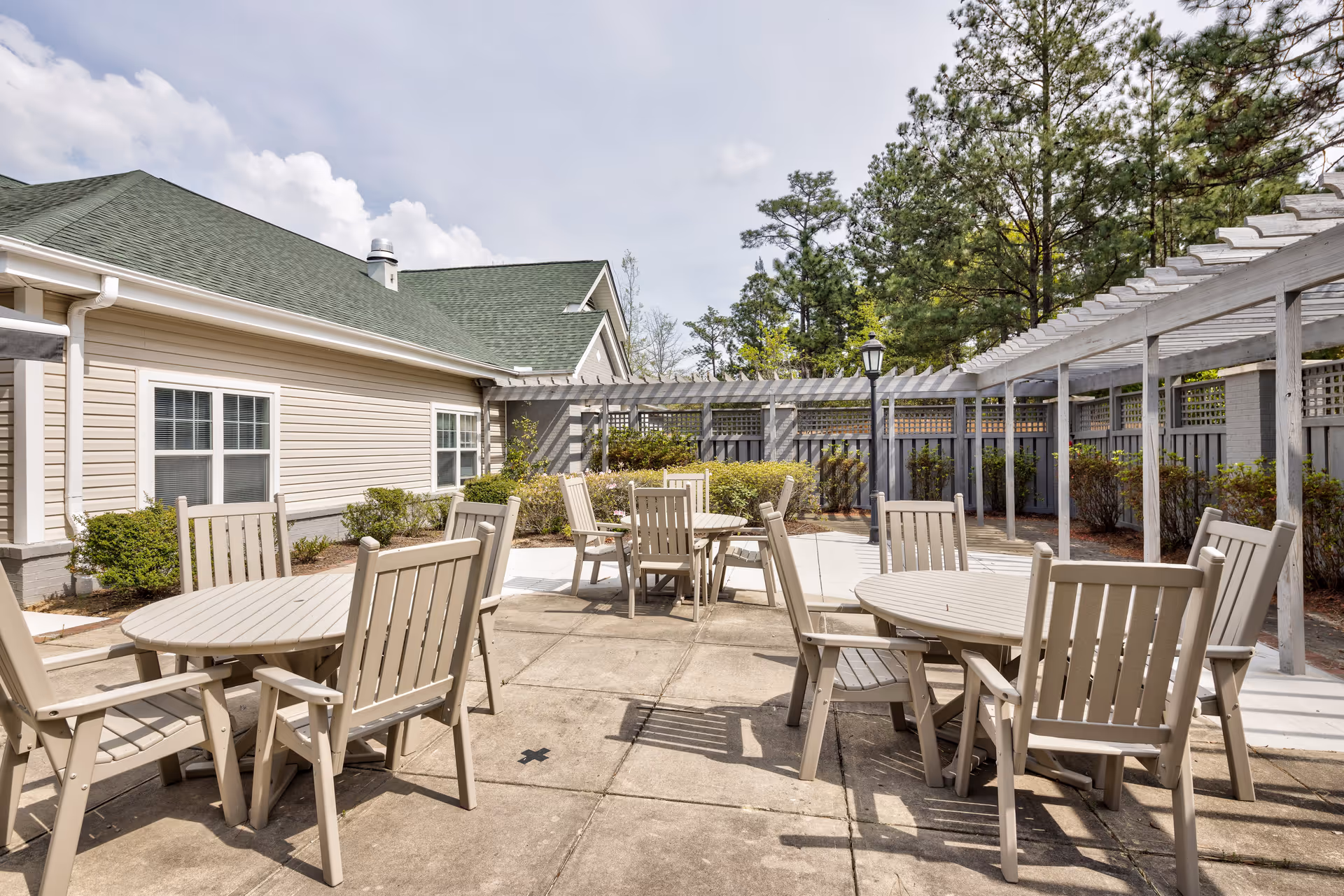 Outdoor patio area with several round tables and wooden chairs arranged on a concrete surface. The patio is adjacent to a beige building with green roofing and surrounded by greenery and trees. A white pergola structure runs along one side of the patio.