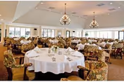 Spacious dining room with multiple round tables covered in white tablecloths, each set with napkins and small floral centerpieces. The room features large windows allowing natural light, floral upholstered chairs, and elegant chandeliers hanging from the ceiling.