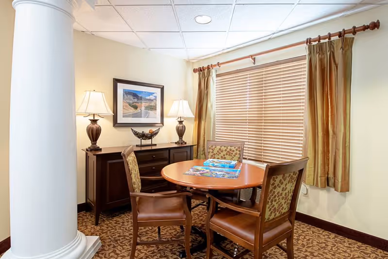 A cozy dining nook featuring a round wooden table with four upholstered chairs, a sideboard with lamps and framed art, and a window with blinds.