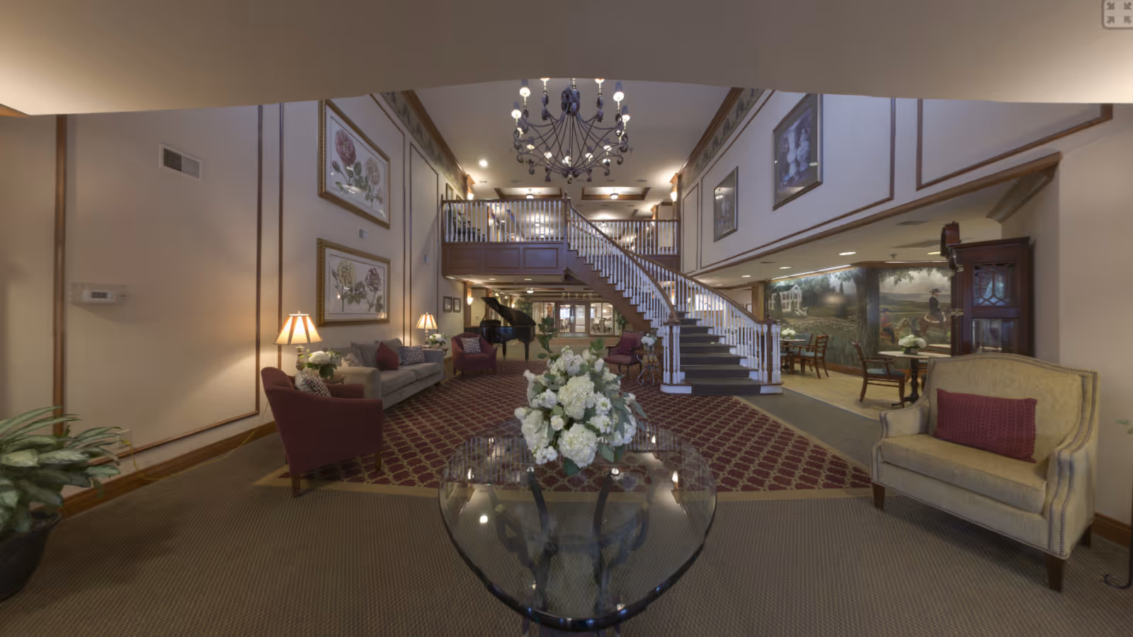 Spacious and elegant senior living facility common area with a large staircase leading to an upper level. The room features comfortable seating including armchairs and sofas, a grand piano, decorative lamps, framed floral artwork on the walls, a chandelier, and a glass table with a flower arrangement in the foreground. There is also a mural on the far wall and a grandfather clock near a small dining area.