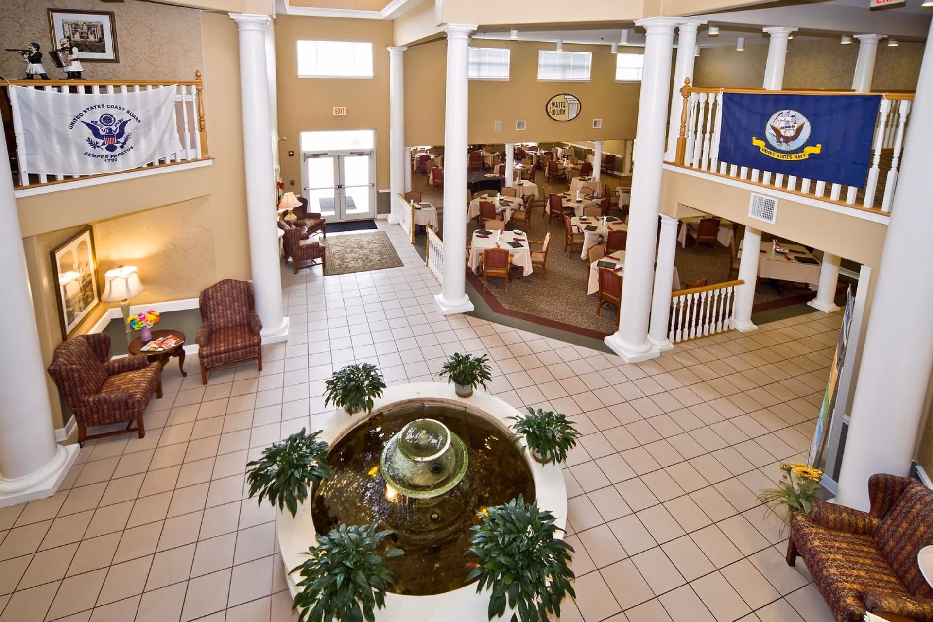 Interior view of The Legacy of Anderson showing a spacious lobby area with a central water fountain surrounded by green plants. The lobby features tiled flooring, white columns, and comfortable seating areas with armchairs and small tables. In the background, there is a dining area with multiple tables covered with white tablecloths and chairs. Flags of the United States Coast Guard and United States Navy are displayed on the upper railings.