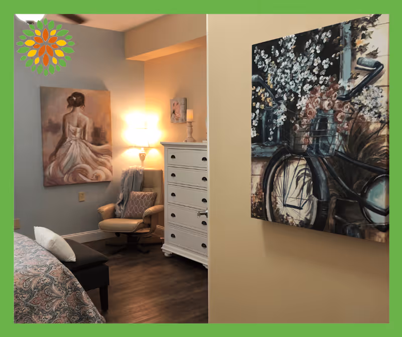 A cozy bedroom corner featuring a beige armchair with a patterned pillow and a throw blanket, a white chest of drawers with a lamp and candle on top, and two paintings on the walls. One painting shows a woman in a flowing dress from the back, and the other depicts a bicycle with flowers in its basket.