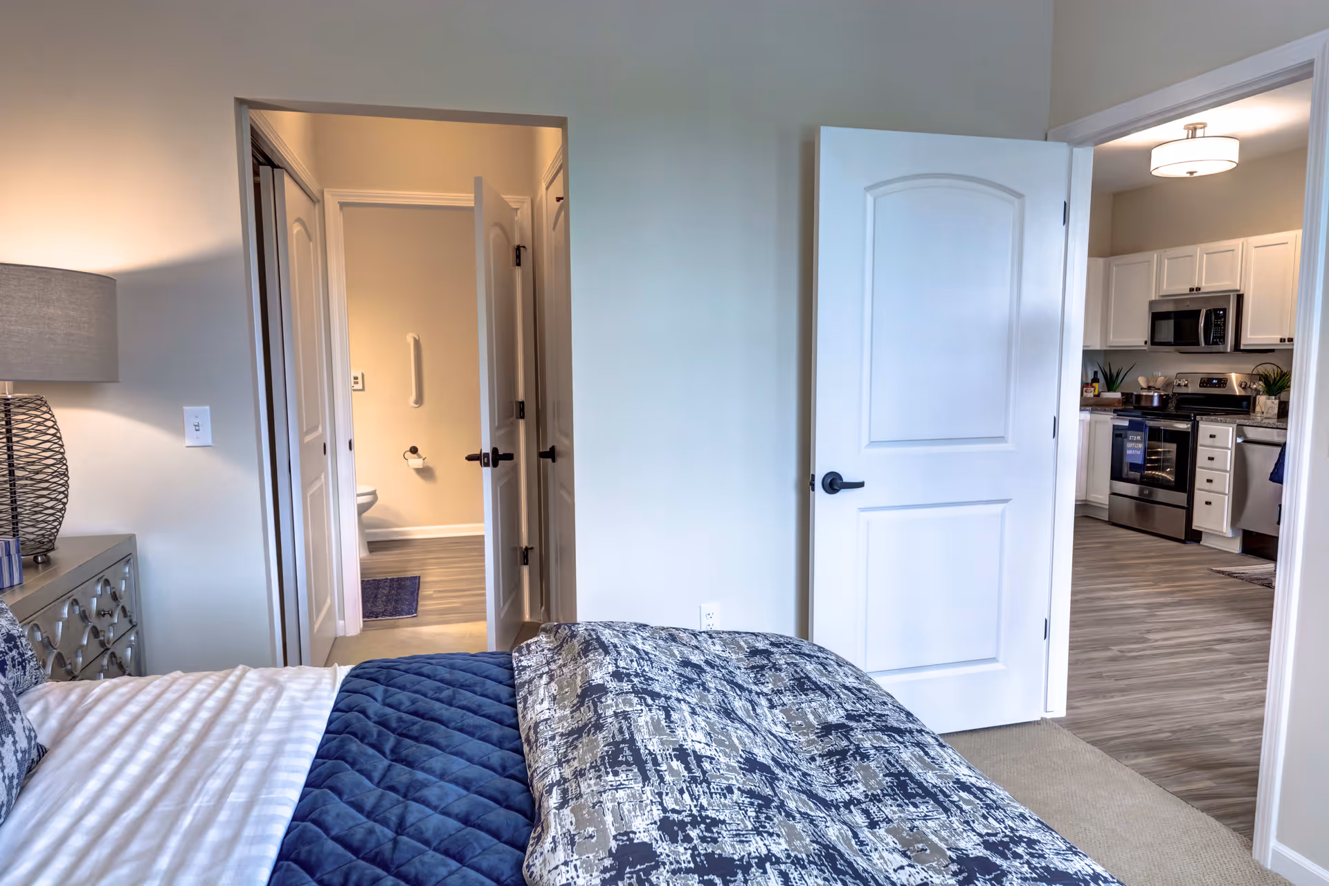 View of a bedroom with a bed covered in a patterned blue and white comforter. To the left, there is a nightstand with a lamp. Two open doors reveal a bathroom with a toilet and a kitchen with white cabinets and stainless steel appliances.