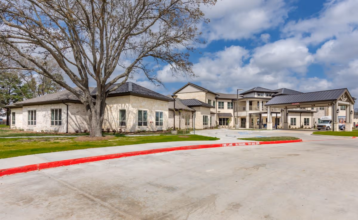 Exterior view of StoneCreek of Copperfield Senior Living facility showing a large, modern building with light-colored stone walls and a dark roof. The building is surrounded by a well-maintained lawn and a large tree with bare branches. The sky is partly cloudy with patches of blue. There is a driveway with a red-painted curb in front of the building.