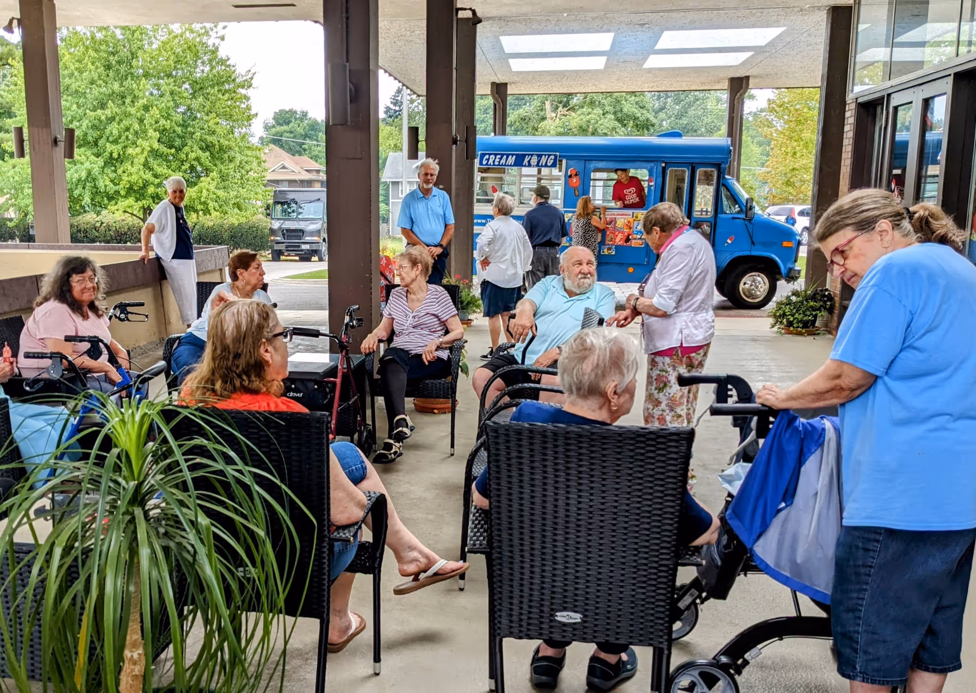 A group of elderly people sitting and standing under a covered outdoor area at Plum Landing Retirement Community. Some are seated on wicker chairs, others are using walkers. In the background, there is a blue ice cream truck with a vendor serving customers. Trees and houses are visible beyond the covered area.