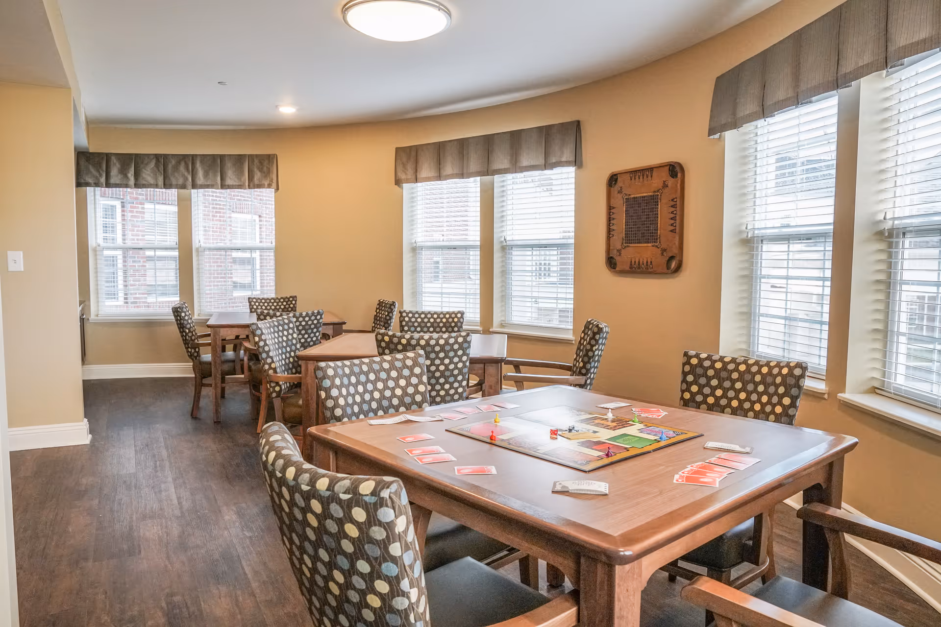 A bright room with several wooden tables and chairs featuring polka dot upholstery. One table in the foreground has a board game set up with cards and game pieces. The room has large windows with blinds and beige walls, creating a cozy and inviting atmosphere.