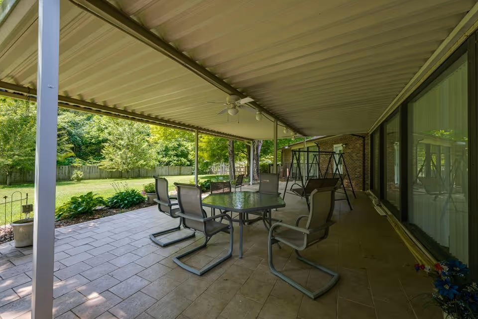 Covered outdoor patio area with a glass-top table and six chairs, a swing set, and a view of a green backyard with trees and a wooden fence.