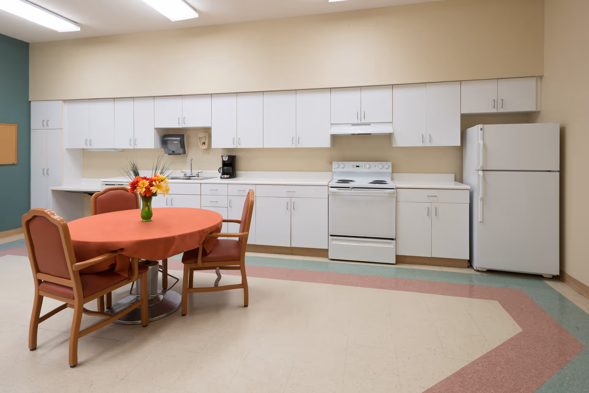 Communal kitchen area with white cabinets, a stove and refrigerator, and a round table with chairs topped by a vase of flowers.