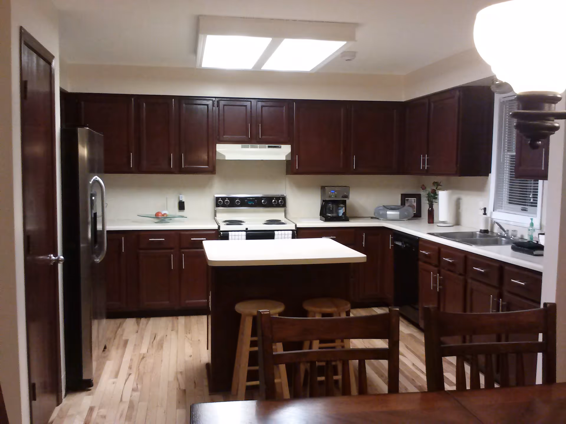 A modern kitchen with dark wood cabinets, a white countertop island with two wooden stools, a stainless steel refrigerator, an electric stove with a range hood, a coffee maker, a double sink under a window with blinds, and hardwood flooring.