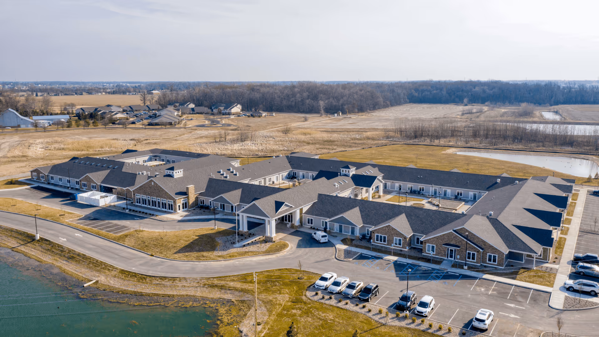 Aerial view of Cedarhurst Senior Living of Fort Wayne, showing a large single-story building with multiple wings, surrounded by parking lots, grassy areas, and a pond. The facility is situated in a rural area with open fields and some trees in the background.
