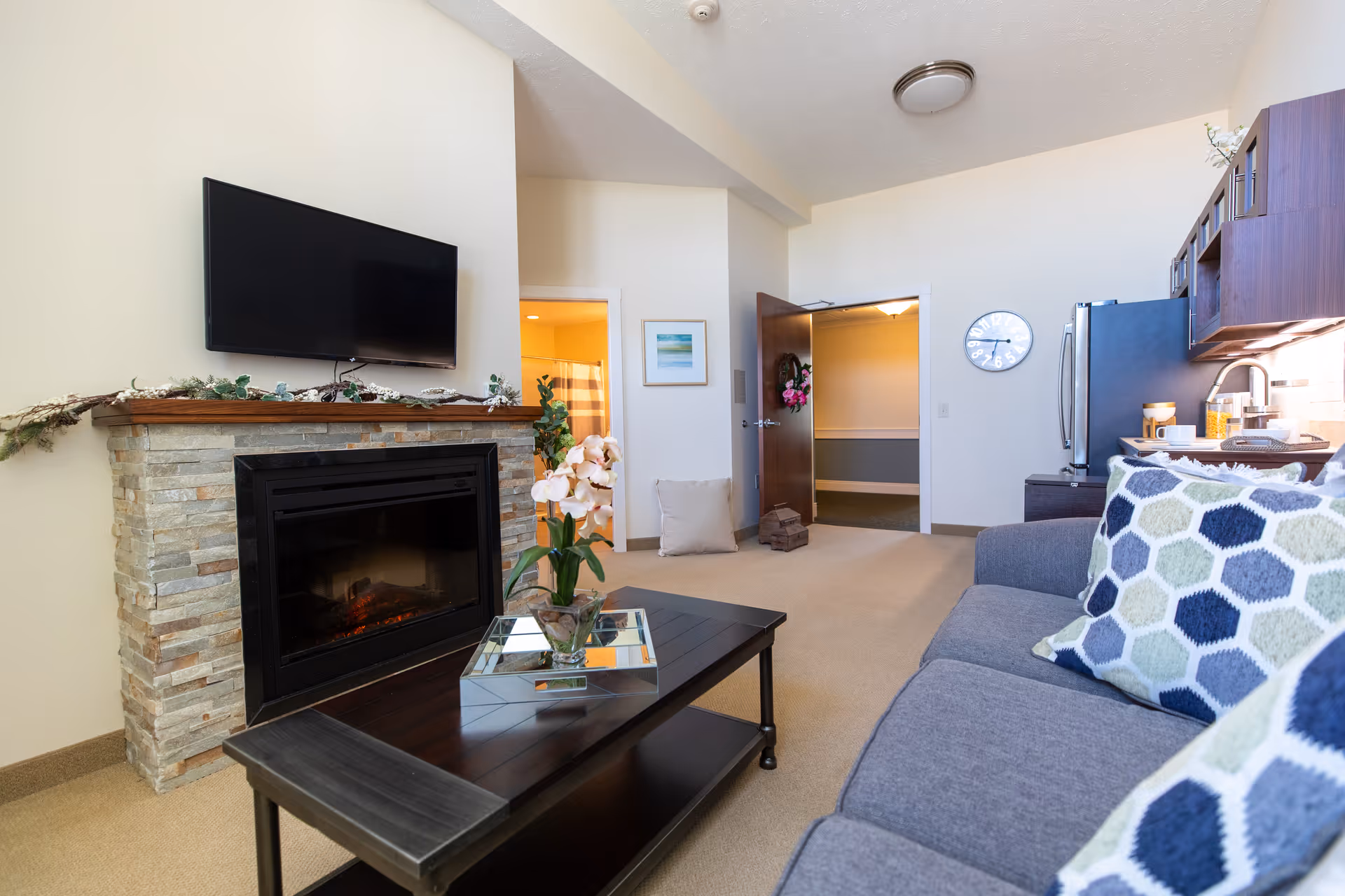 A cozy living room area with a stone fireplace and a mounted flat-screen TV above it. A dark wooden coffee table with a glass vase holding flowers sits in front of a gray sofa adorned with patterned cushions. The room has beige walls and carpet, with an open doorway leading to a bathroom and another open door leading to a hallway. A clock is mounted on the wall near a stainless steel refrigerator and kitchen cabinets.