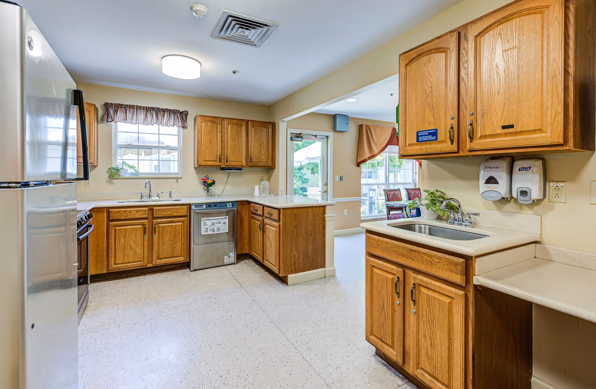 A bright kitchen area with wooden cabinets, a stainless steel dishwasher, a refrigerator, and a stove. There is a window with a valance above the sink, and a small plant and flowers on the counter. The kitchen opens into a room with chairs and large windows letting in natural light.