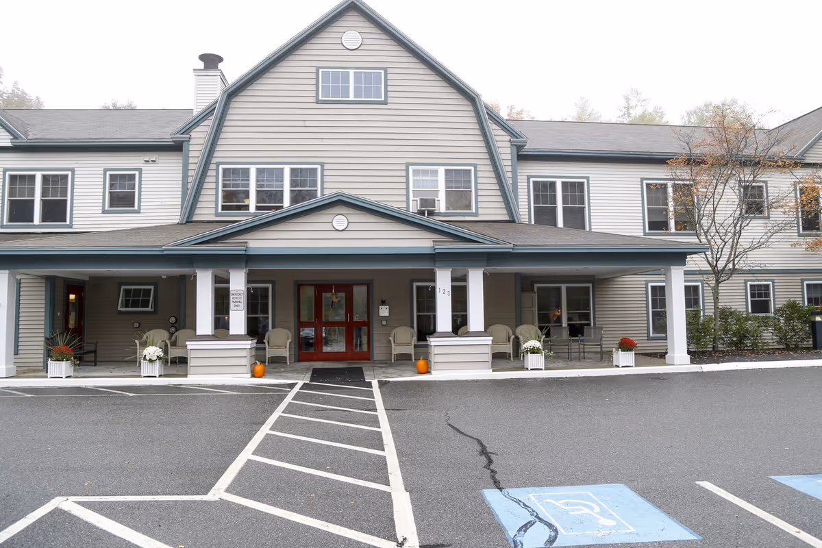Exterior front entrance of a two-story senior living building with a covered porch, red double doors, chairs, planters, and marked handicap parking spaces.