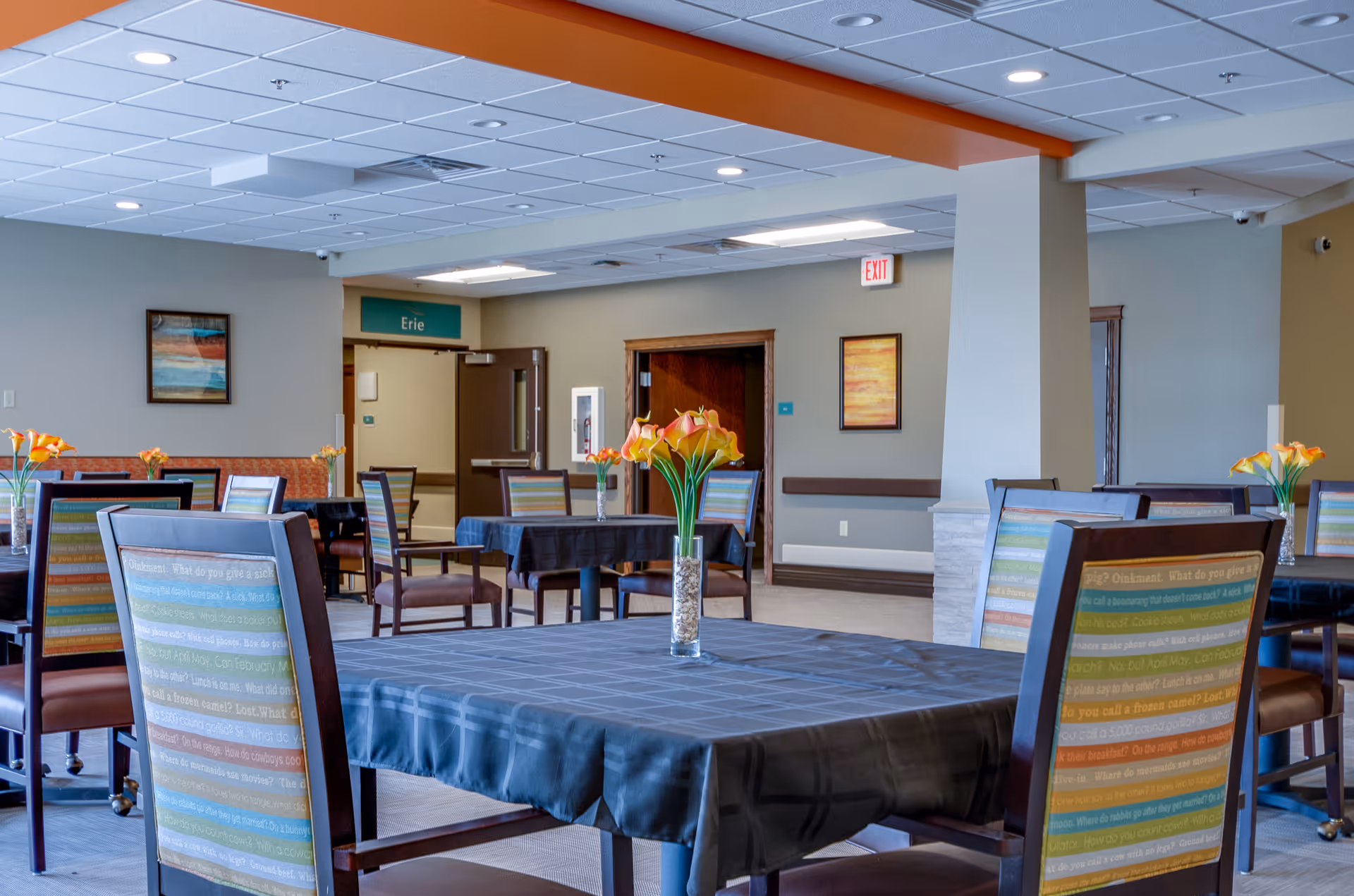 Bright dining room with multiple tables covered by dark tablecloths, striped chairs, vases of flowers, and a doorway sign reading 'Erie'.