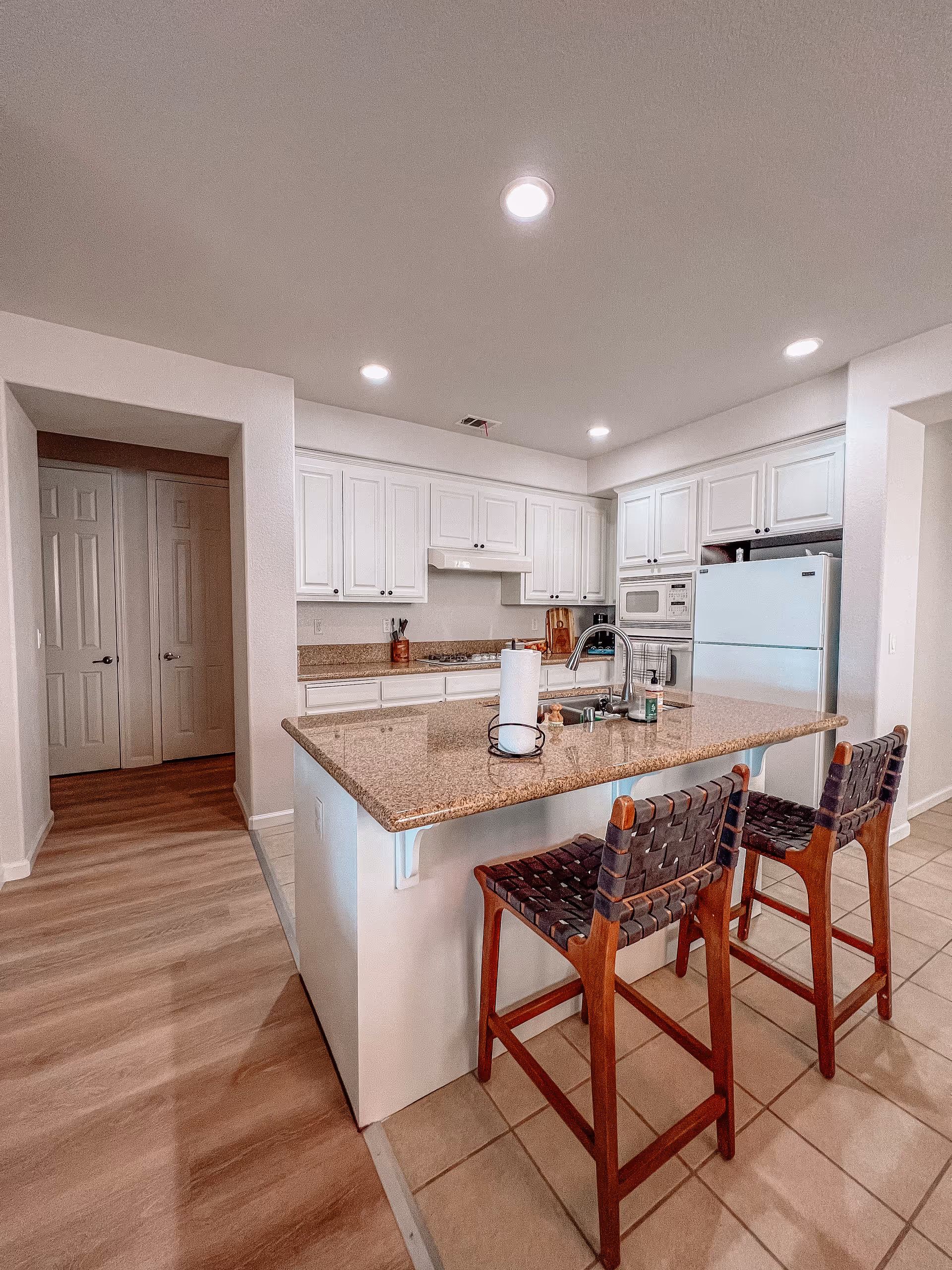A modern kitchen with white cabinets, a granite countertop island with a sink, and two wooden bar stools with woven seats. The kitchen features built-in appliances including a microwave and refrigerator. The floor is a combination of tile and wood, and recessed ceiling lights illuminate the space.