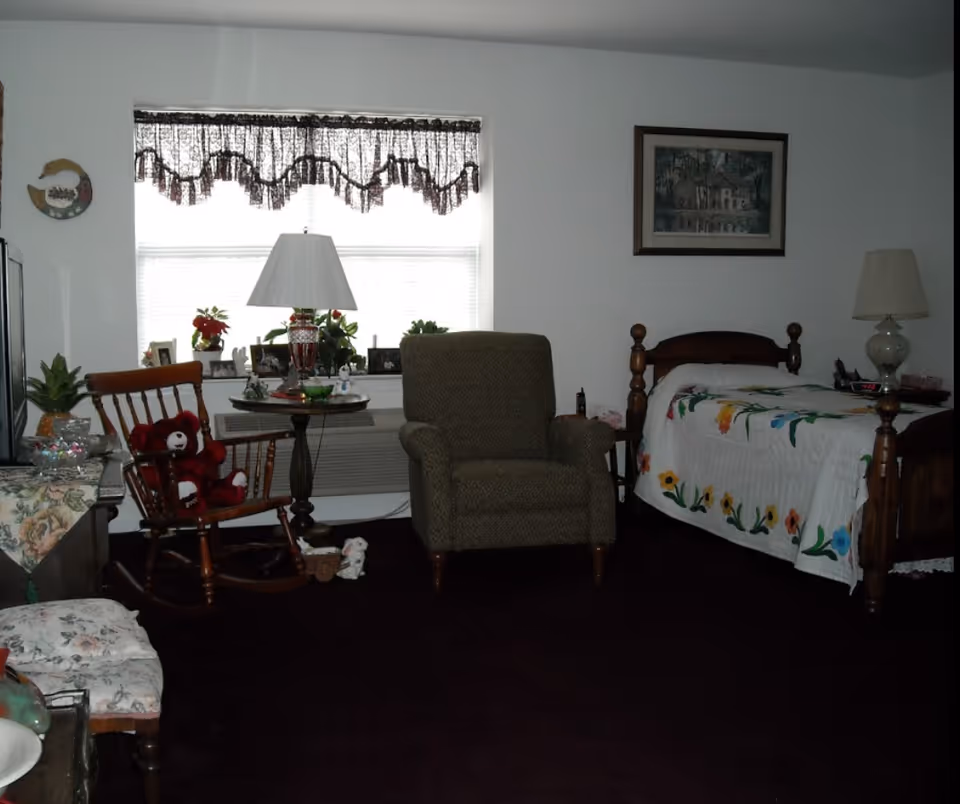 A cozy bedroom in an assisted living facility featuring a wooden bed with a colorful floral quilt, a cushioned armchair, a wooden rocking chair with a red teddy bear, a small round table with a lamp and plants, and a window with lace curtains letting in natural light.