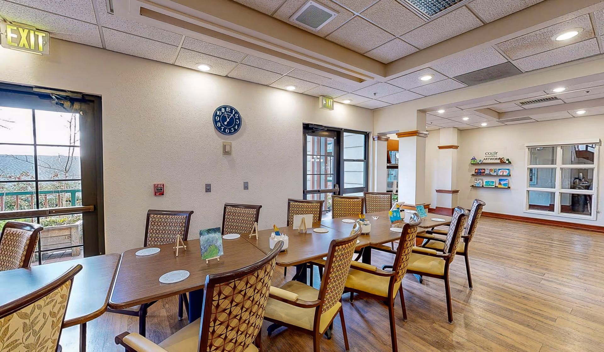 Communal dining/activity room with a long table surrounded by chairs, a wall clock, and exit doors.
