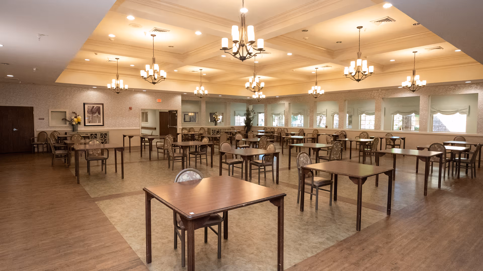 A spacious dining room with multiple wooden tables and chairs arranged neatly. The room features a coffered ceiling with several chandeliers providing warm lighting. Large windows with valances allow natural light to enter, and the walls are decorated with framed artwork and floral arrangements.