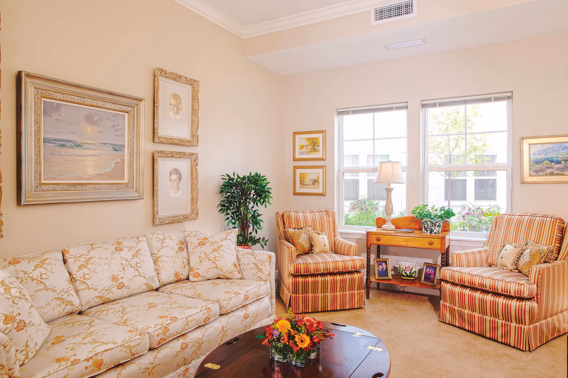 A cozy living room with a floral patterned sofa, two striped armchairs, a wooden coffee table with a flower arrangement, and a wooden side table with a lamp and framed photos. The walls are decorated with framed paintings and portraits, and two large windows let in natural light, showing greenery outside.