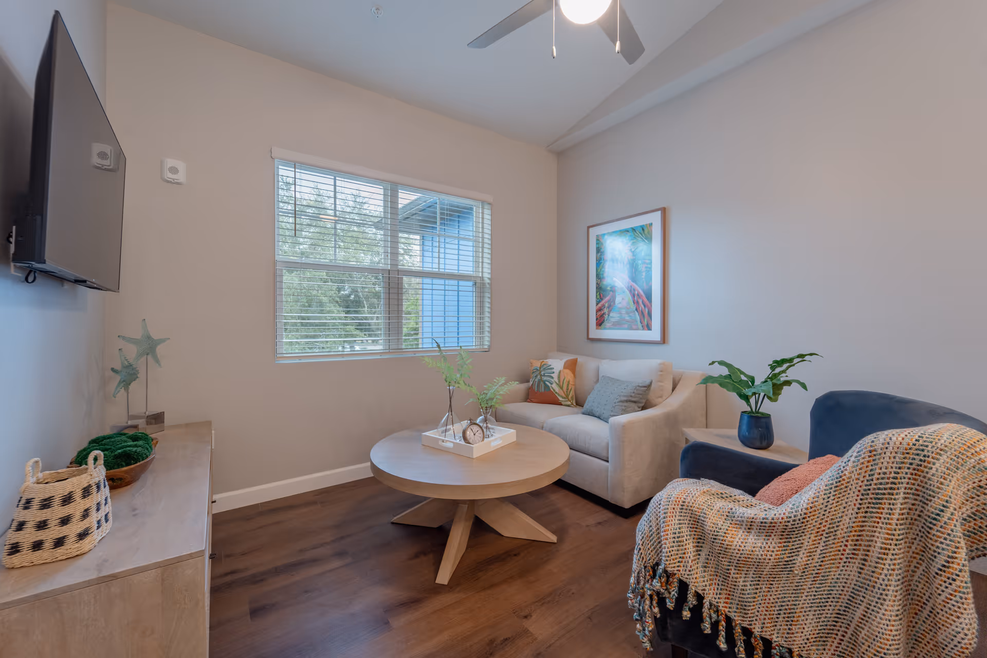 A cozy living room with a beige sofa adorned with colorful cushions, a round wooden coffee table with decorative plants and a clock, a blue armchair draped with a multicolored throw blanket, a wooden sideboard with decorative items, a wall-mounted flat-screen TV, a window with blinds letting in natural light, and a ceiling fan with a light.