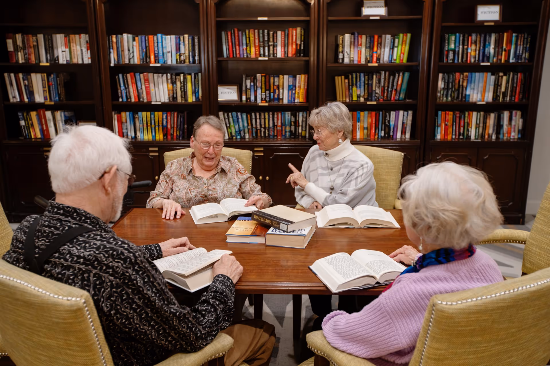 Four elderly individuals sitting around a wooden table in a library or reading room, each with an open book. Behind them are dark wooden bookshelves filled with books, labeled 'Fiction'. The group appears engaged in reading or discussion.