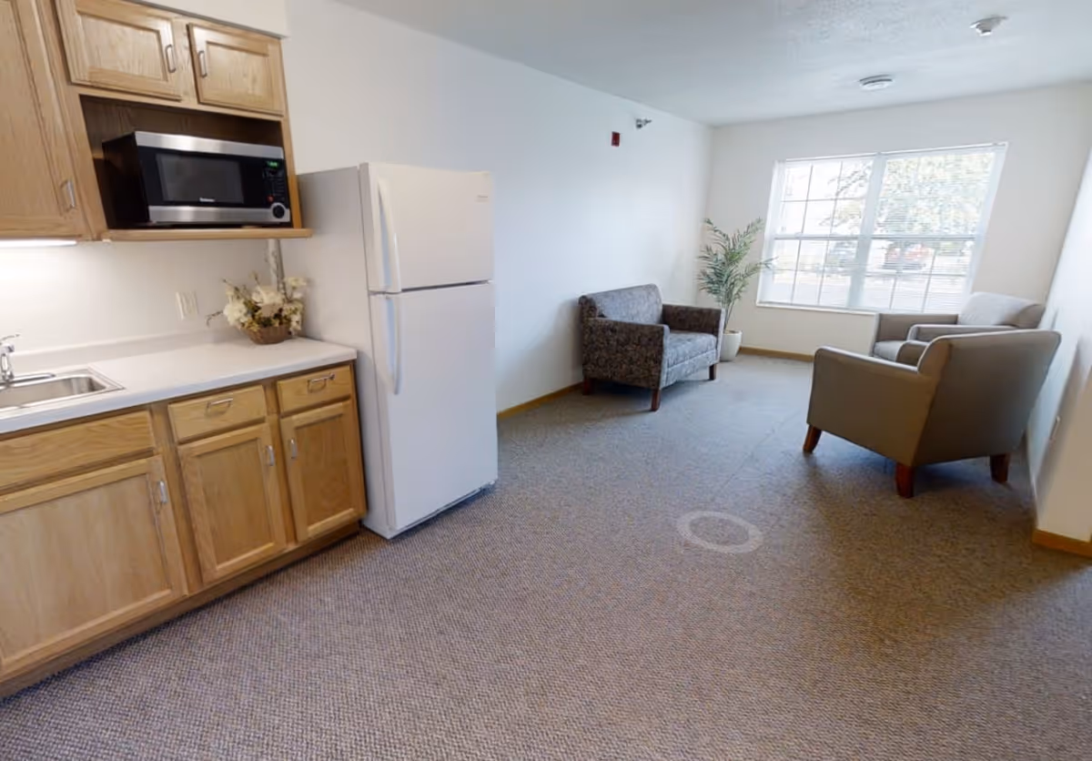 A small living area with two armchairs and a loveseat arranged near a large window with blinds. To the left, there is a kitchenette with wooden cabinets, a white refrigerator, a microwave, a sink, and a small flower arrangement on the counter. The room has carpeted flooring and white walls.