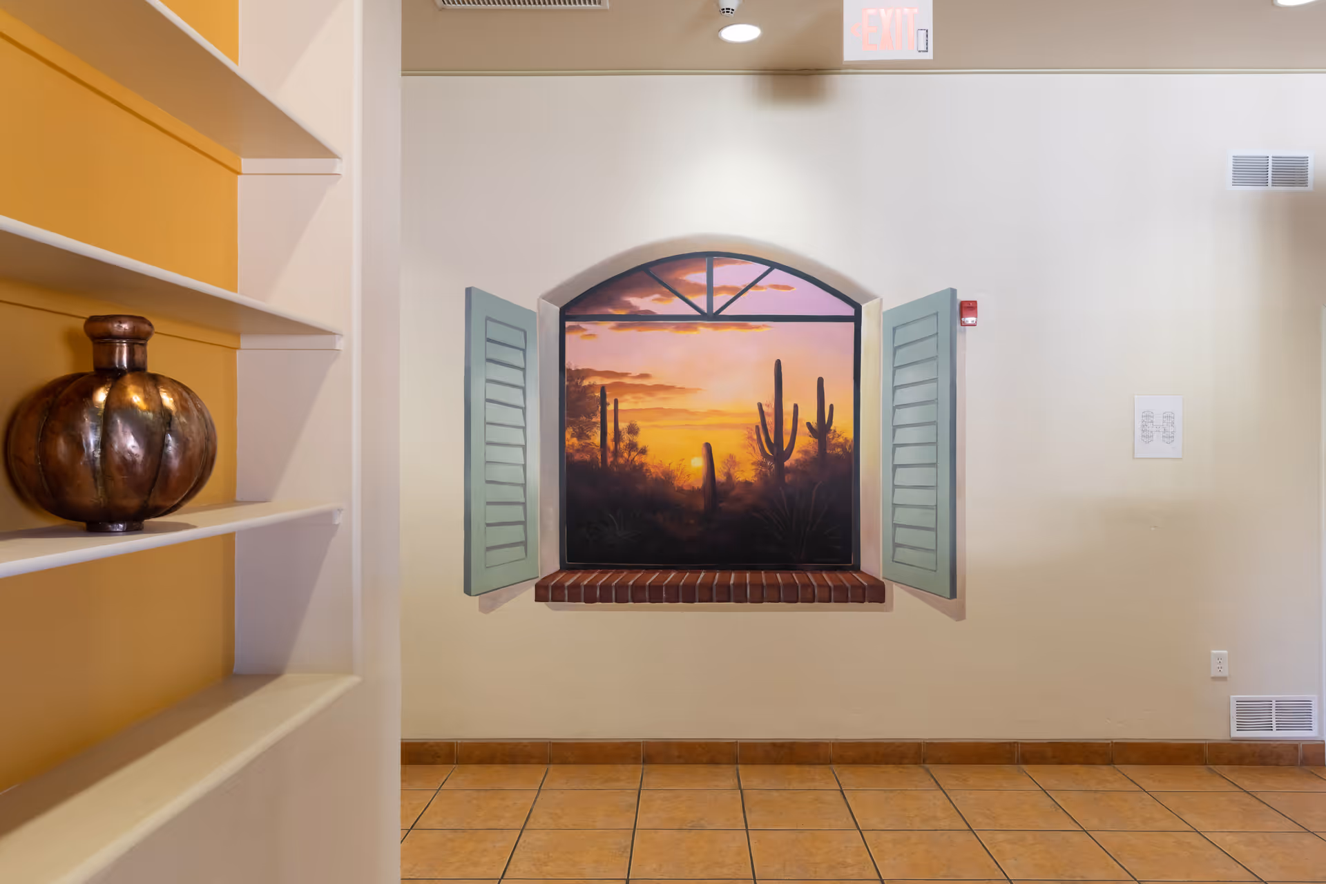 Interior wall with a decorative window frame featuring open green shutters and a painted desert sunset scene with cacti. To the left, built-in shelves with a bronze decorative vase. The floor is tiled and the walls are light-colored.