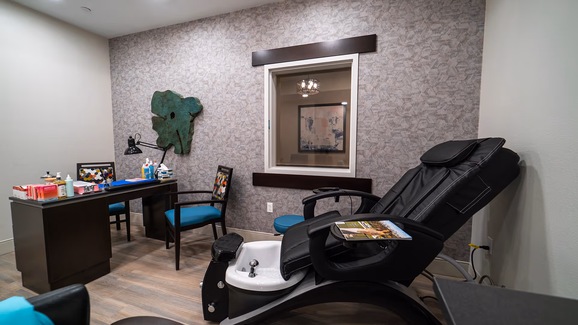 A pedicure station in a senior living facility featuring a black reclining chair with a foot bath attached. Next to it is a dark wooden desk with two chairs, various nail care products, and a desk lamp. The wall behind has a textured wallpaper with a green abstract wall decoration and a window showing a framed artwork and a chandelier in the adjacent room.