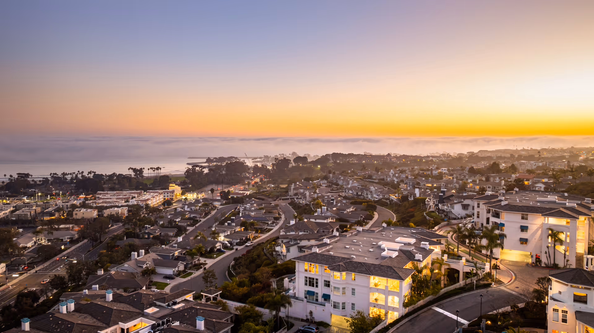 Aerial view of a coastal residential area at sunset with buildings, roads, palm trees, and the ocean in the background under a colorful sky.