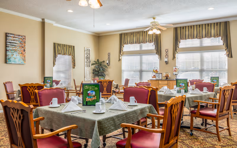A bright dining room with multiple tables covered in green tablecloths, each set with white napkins folded in a triangular shape, white cups, and silverware. The chairs have wooden frames with red cushioned seats and backs. Large windows with blinds and valances let in natural light. The room features ceiling fans with lights, a patterned carpet, and decorative wall art and plants.