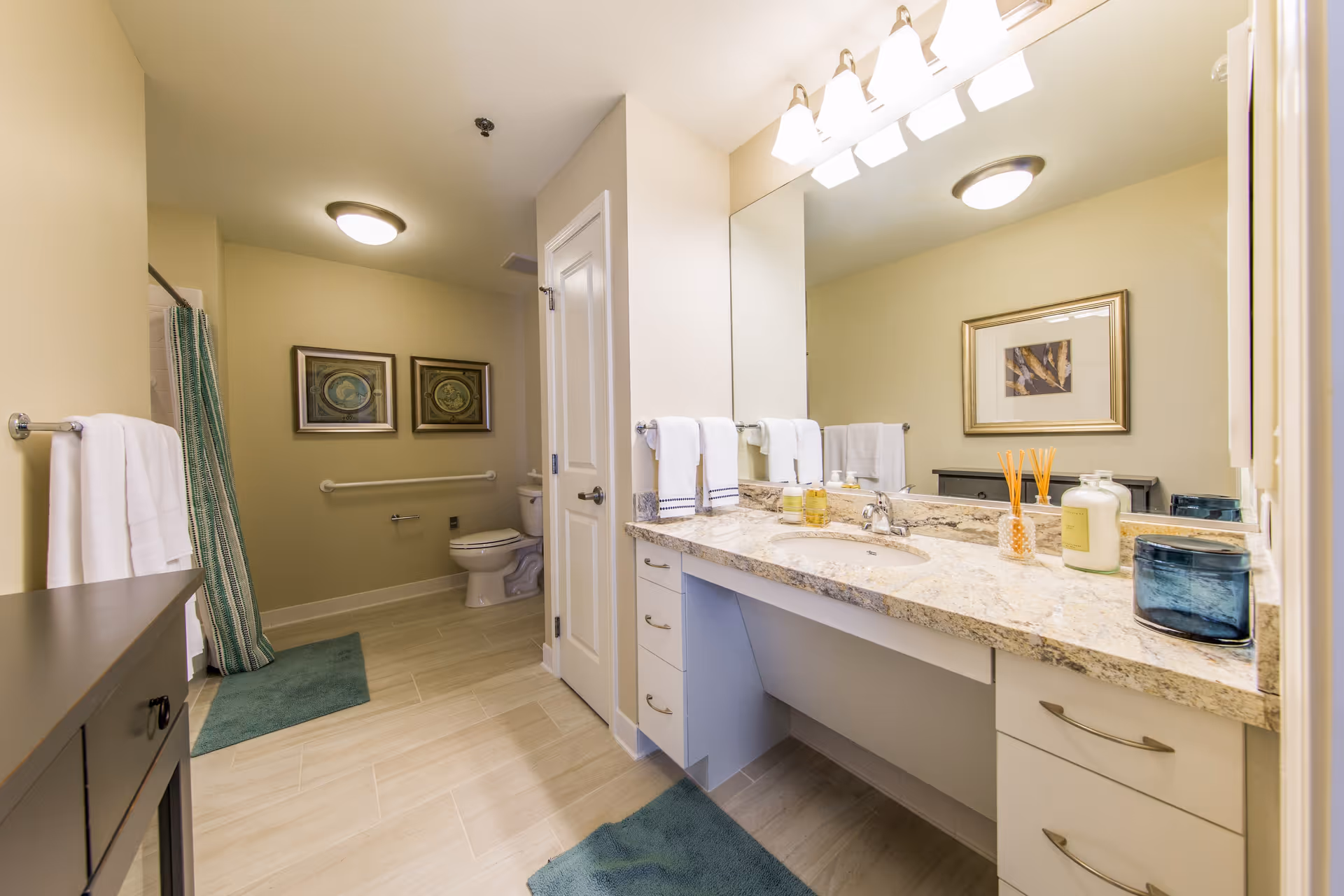 A spacious bathroom with a large granite countertop featuring a sink, toiletries, and decorative items. There is a large mirror above the countertop with multiple light fixtures. The bathroom has light-colored walls and flooring, a toilet in the background, a shower with a green and white striped curtain, two framed pictures on the wall, and several blue bath mats on the floor.