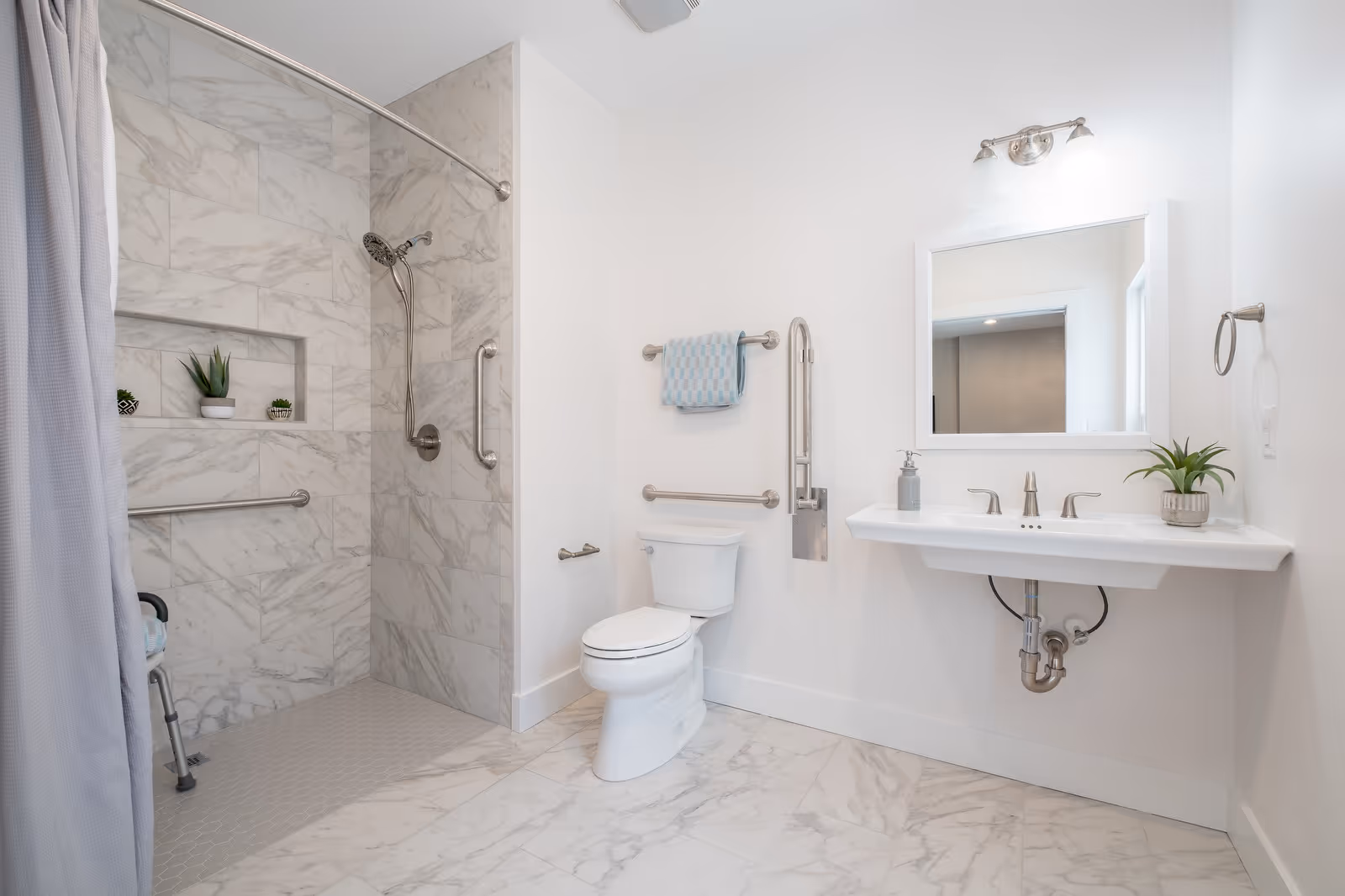 A modern, accessible bathroom with white marble tile flooring and walls. The bathroom features a walk-in shower with a curved shower rod and curtain, grab bars, and a built-in shelf with small potted plants. There is a white toilet with grab bars on the wall, a white wall-mounted sink with a silver faucet, a soap dispenser, and a small potted plant. Above the sink is a rectangular mirror with a light fixture. The walls are painted white, and the bathroom has a clean, bright appearance.