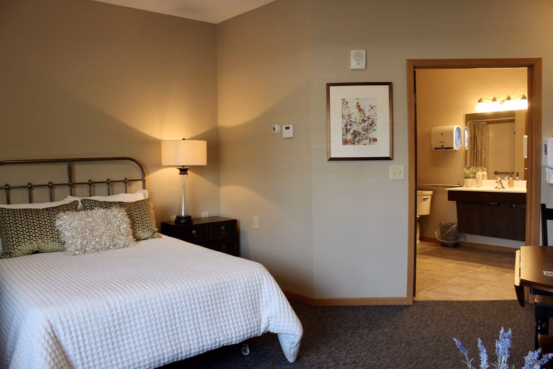 A furnished bedroom with a made bed, bedside lamp, framed artwork, and an open doorway leading to a bathroom.