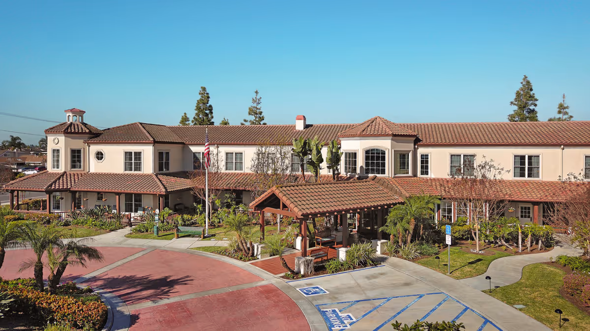 Exterior view of a two-story senior living facility with a tiled roof, beige walls, and multiple windows. The entrance features a covered porch with wooden beams and a small garden area with palm trees and other plants. There is a circular driveway with marked parking spaces, including handicapped spots, and an American flag near the entrance.