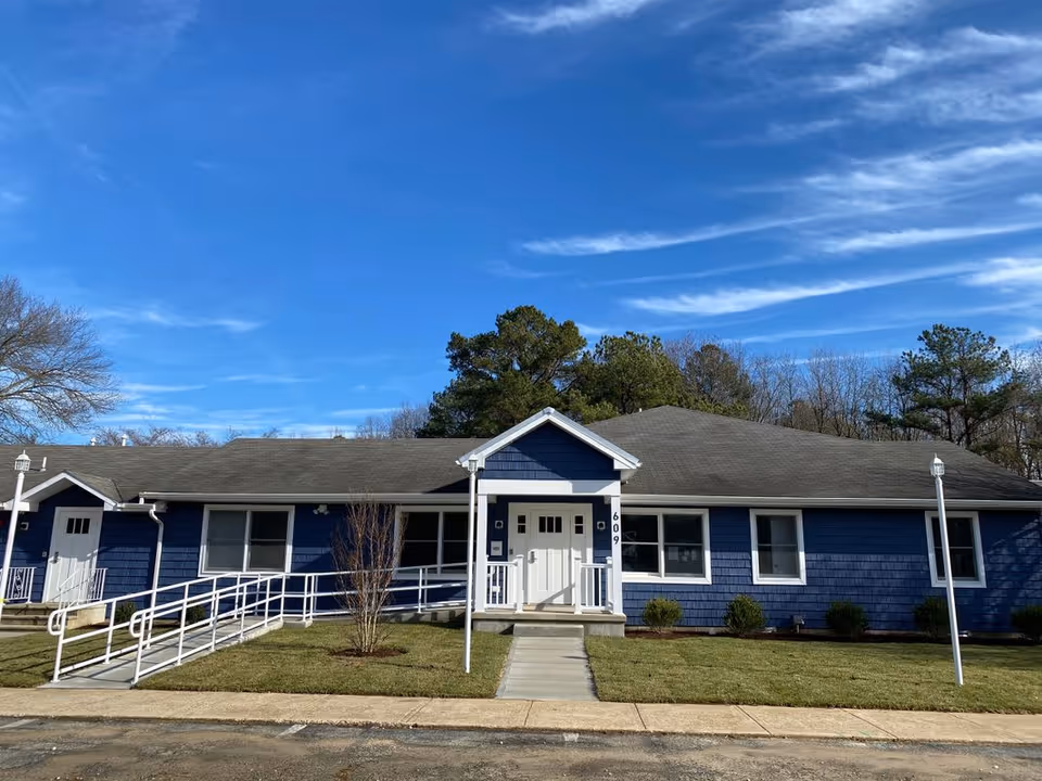 Front exterior view of a single-story blue building with white trim, a gray roof, and a white entrance door with the number 609. There is a concrete walkway leading to the entrance, flanked by a white handrail ramp and small bushes on a grassy lawn. The sky is clear with some wispy clouds and trees are visible in the background.