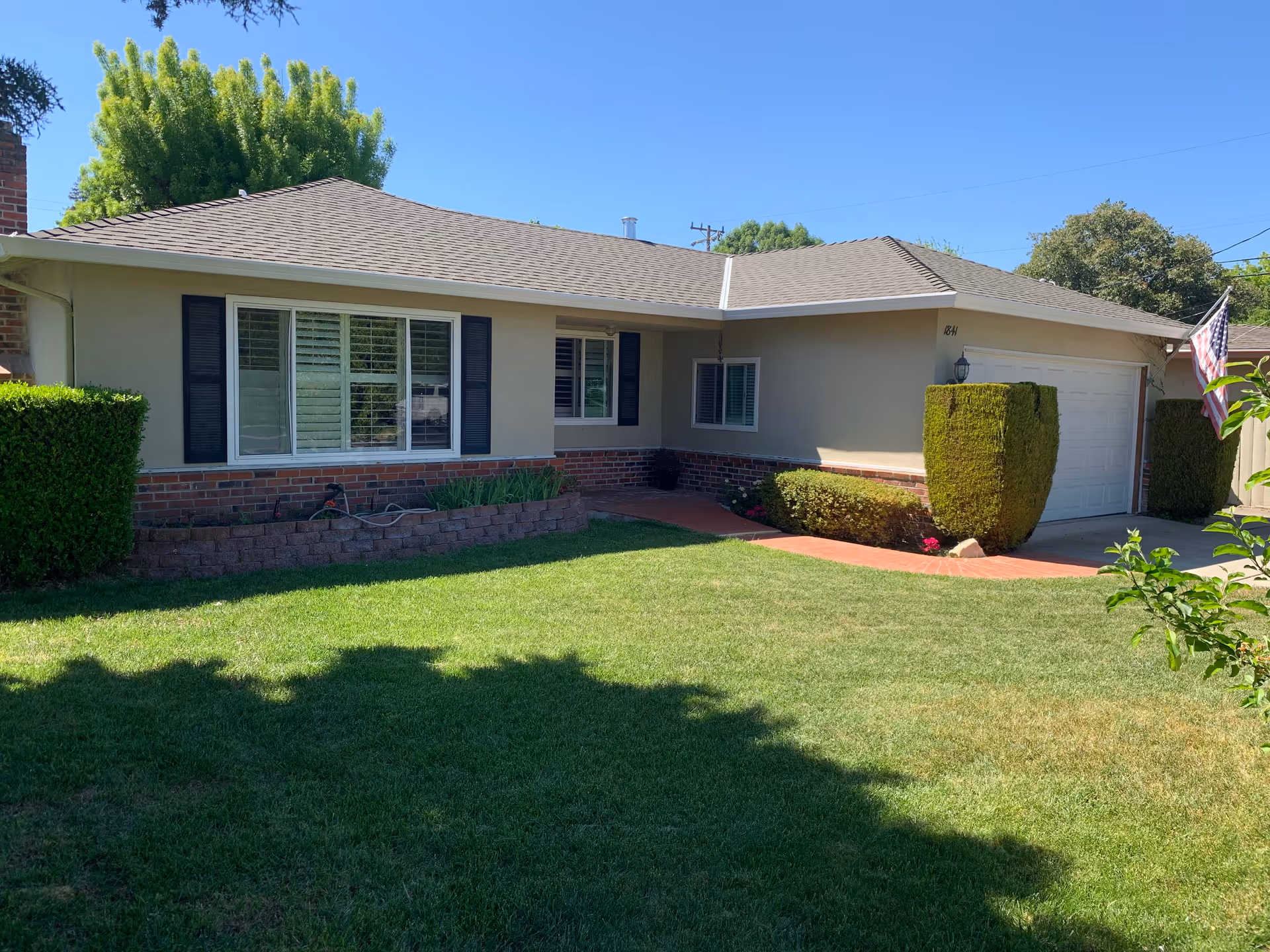 Front exterior of a single-story ranch-style house with a manicured lawn, trimmed hedges, garage, and an American flag.