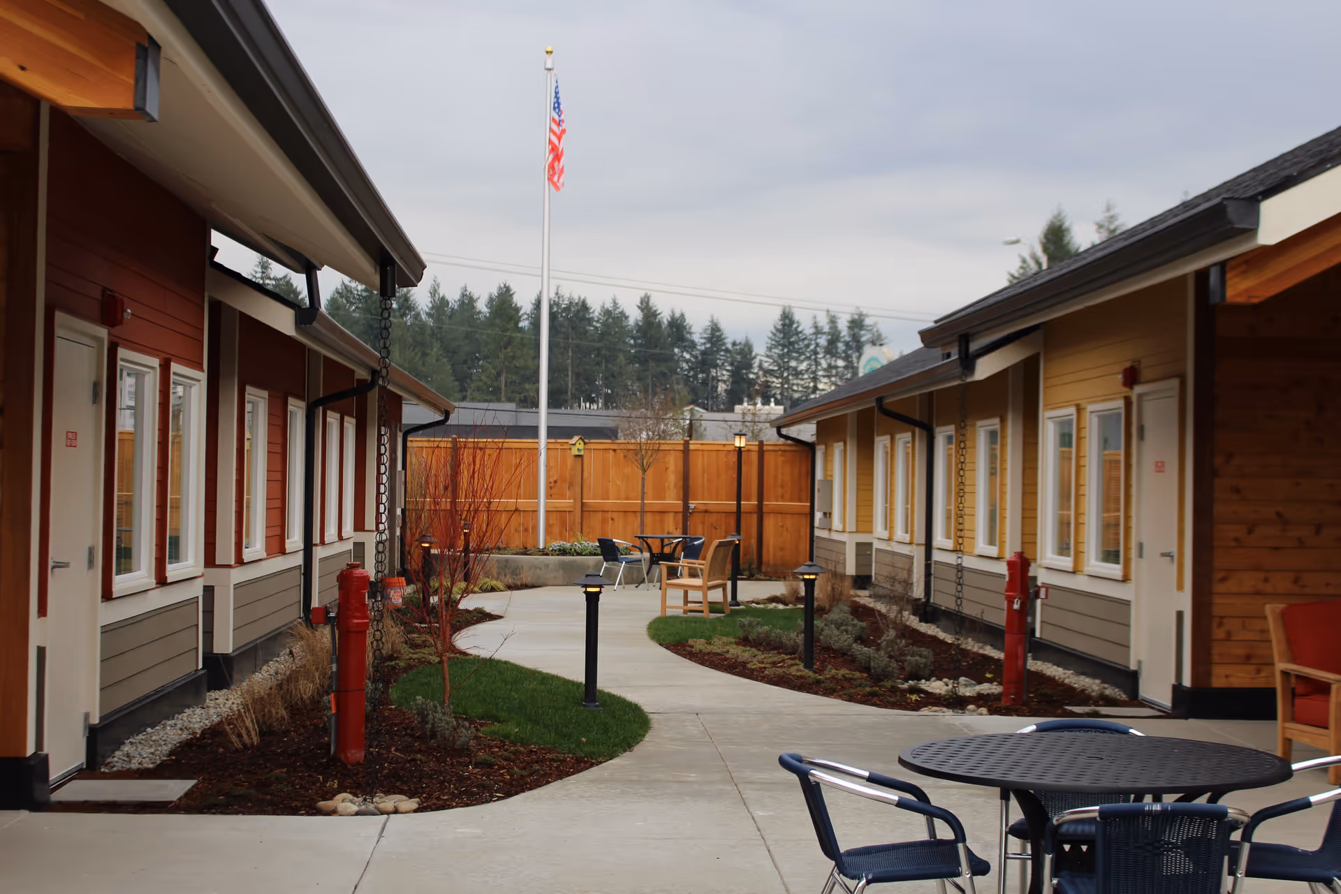 Courtyard between single-story cottages with a paved walkway, outdoor tables and chairs, landscaping, and an American flag.