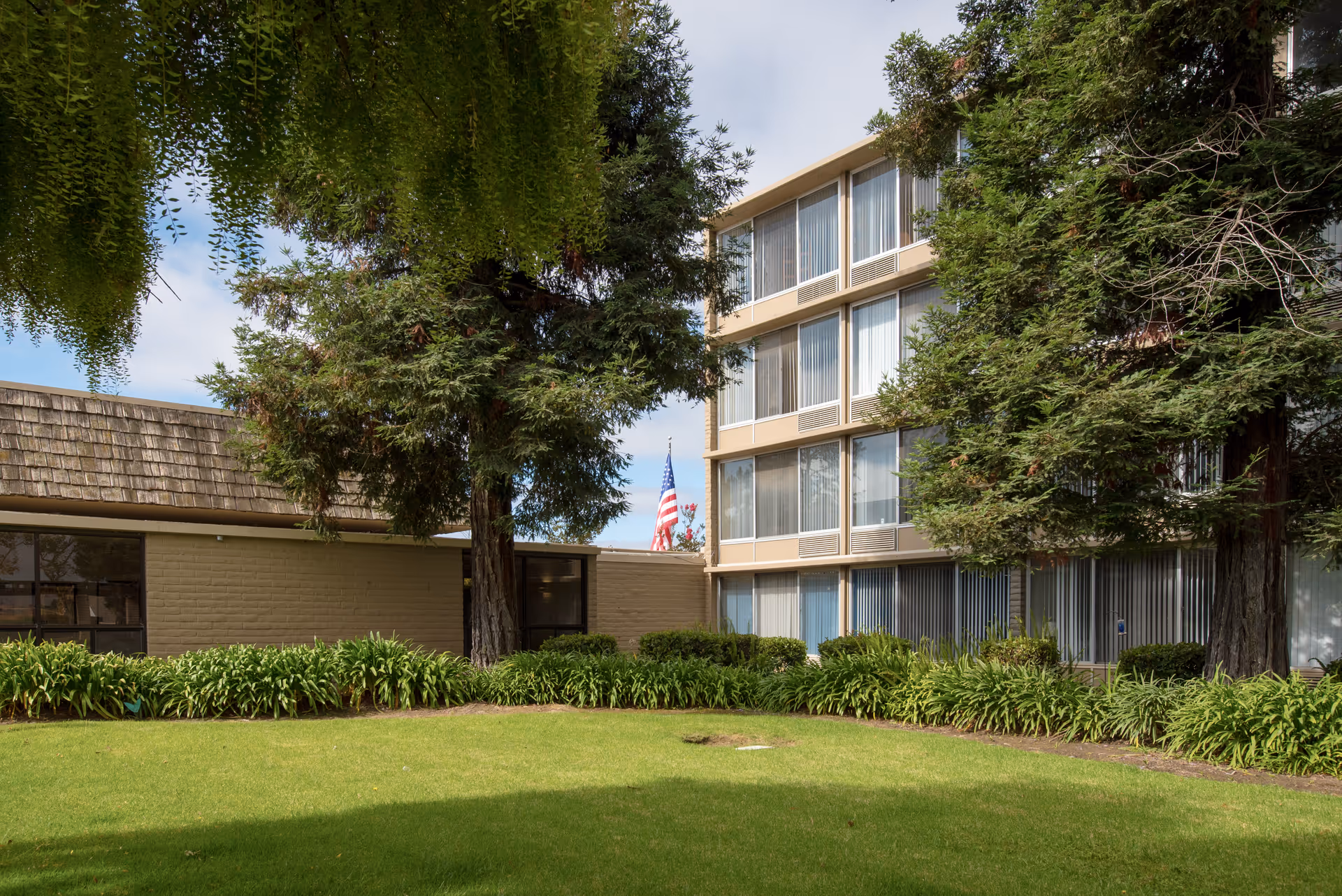 Exterior view of a multi-story senior living building with a lawn, trees, and an American flag in the courtyard.