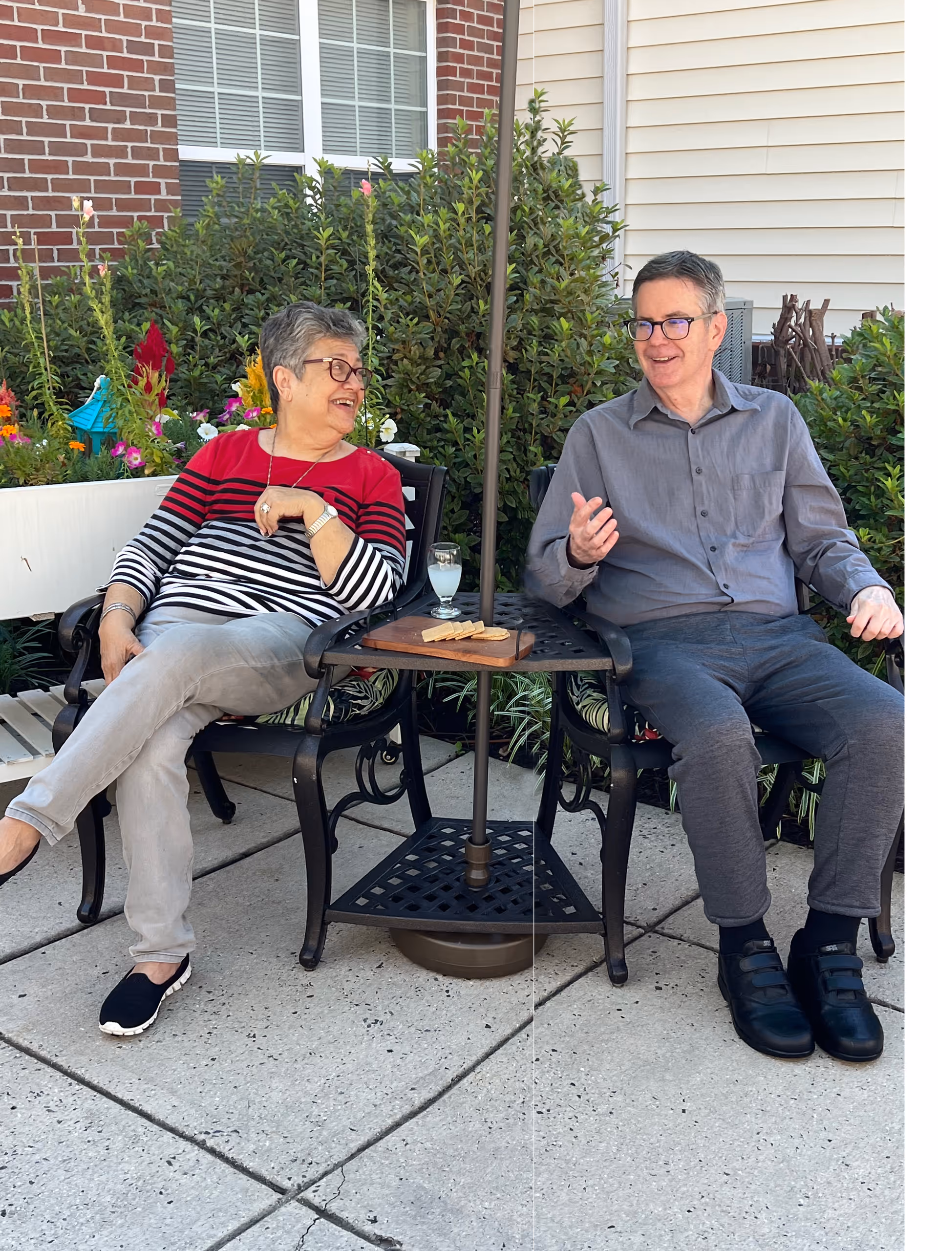 Two elderly people sitting outdoors on black metal chairs with a small table between them holding a glass of water and some crackers. They are engaged in conversation and smiling. Behind them are green bushes and the exterior walls of a building with a window.