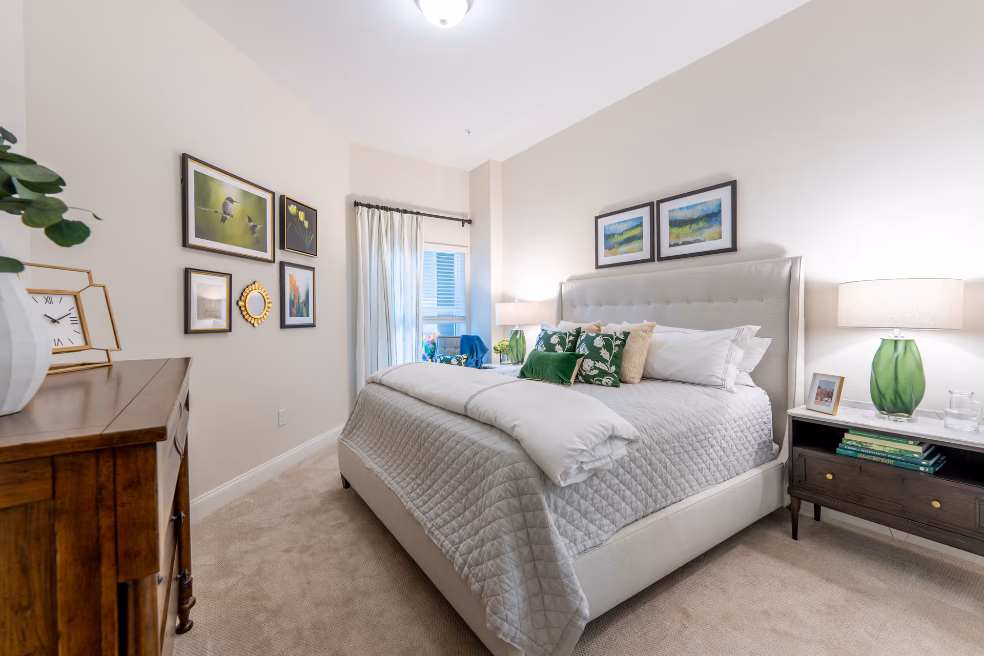 A bright and cozy bedroom featuring a large upholstered bed with a quilted gray bedspread and multiple pillows, two framed landscape paintings above the headboard, a wooden nightstand with a green glass lamp and books, a wooden dresser with a clock and decorative items, and a wall adorned with framed pictures and a small round mirror. A window with white curtains allows natural light into the room.