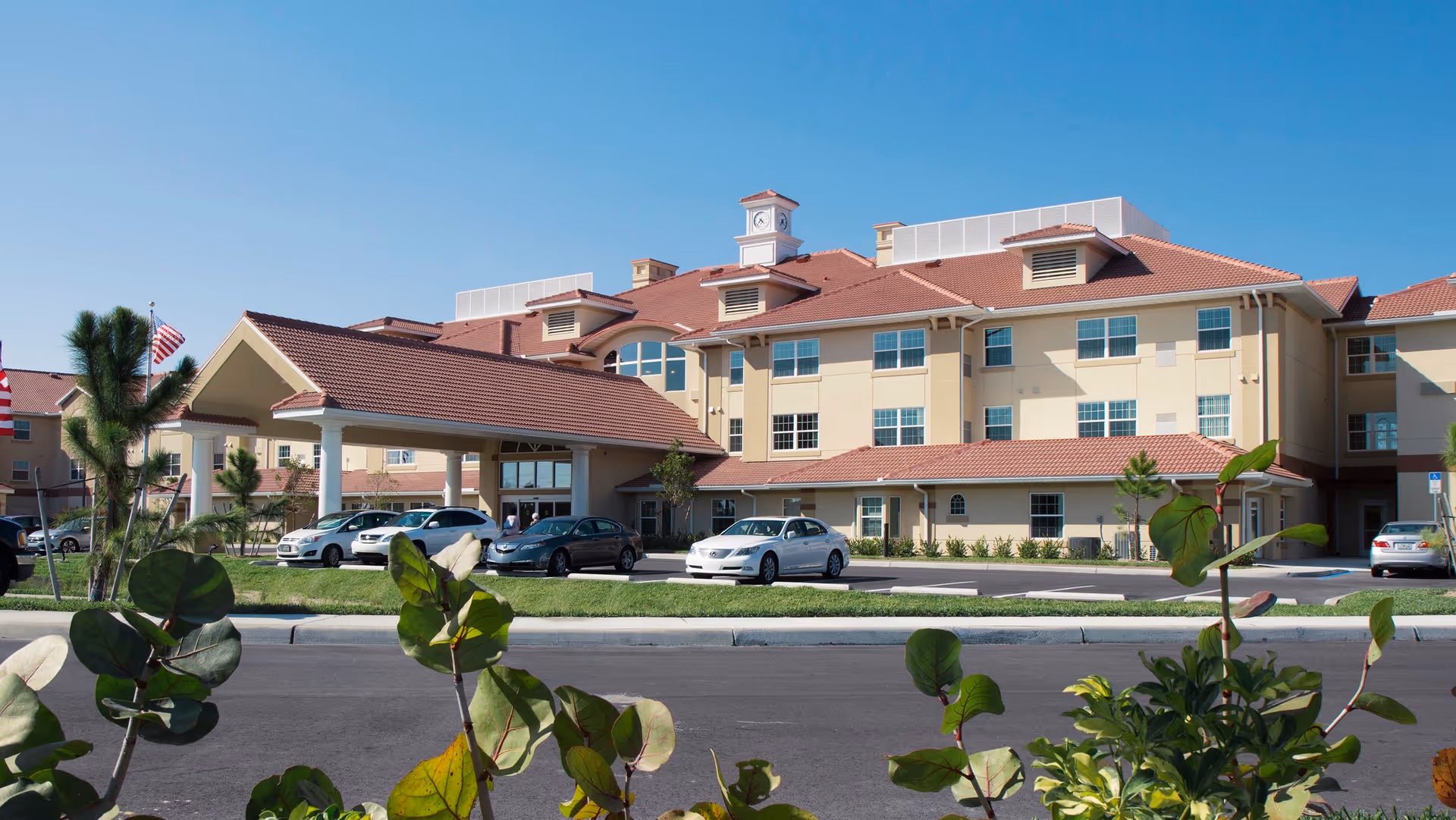 Exterior view of Bradford Square Retirement Community building with a covered entrance, several parked cars, and clear blue sky. Some greenery and small trees are visible in the foreground.