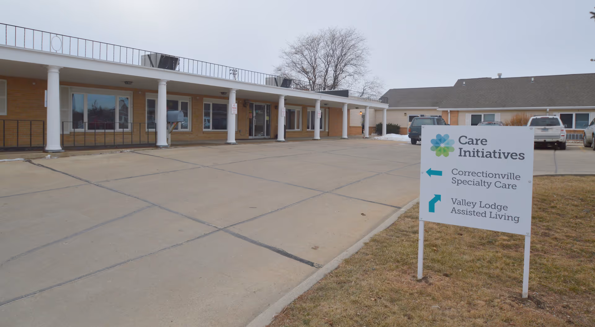Exterior view of a single-story building with a covered entrance supported by white columns. There is a paved driveway in front and a sign on the grass indicating directions to Correctionville Specialty Care and Valley Lodge Assisted Living. Several vehicles are parked near the building.