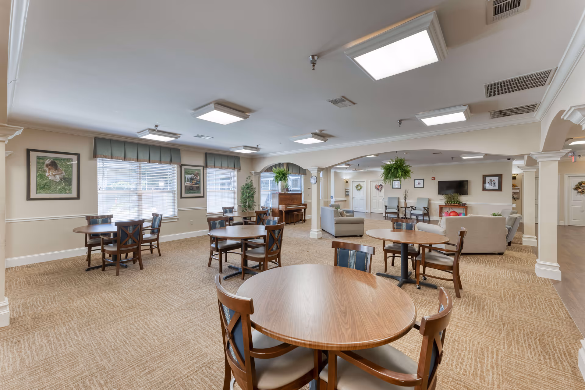 A spacious and well-lit common area in a senior living facility featuring several round wooden tables with chairs, a seating area with sofas and armchairs, framed artwork on the walls, large windows with blinds, and a piano in the corner. The room has beige carpeting and neutral-colored walls with white trim and columns.