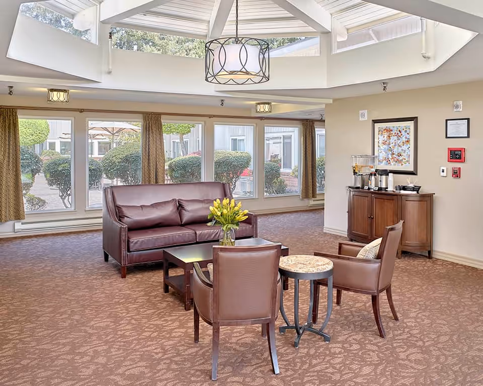 Bright senior living common lounge with leather sofa and chairs around a coffee table and large windows overlooking a landscaped courtyard.