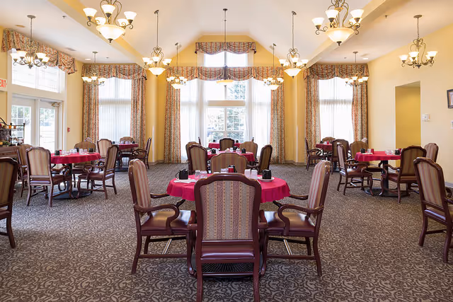 A spacious dining room with multiple round tables covered with red tablecloths, each surrounded by wooden chairs with cushioned seats and backs. Large windows with patterned curtains allow natural light to fill the room. Elegant chandeliers hang from the ceiling, illuminating the space.