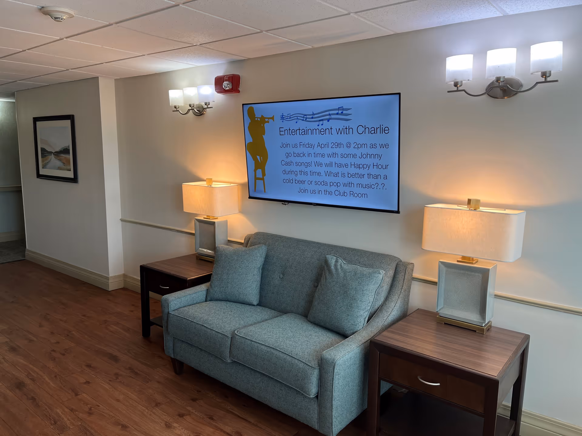 A cozy seating area in a senior living facility with a light blue loveseat flanked by two wooden side tables, each with a lamp. Above the loveseat is a wall-mounted TV screen displaying an announcement for entertainment with Charlie, featuring Johnny Cash songs and a happy hour event. The room has wooden flooring, soft lighting from wall sconces, and a framed landscape painting on the adjacent wall.
