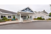 Exterior view of a single-story senior living facility building with a covered entrance, a paved driveway, and landscaped bushes and plants around the building.